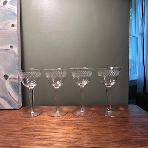 May include: Four clear glass stemmed drinking glasses with a decorative etched design. The glasses are arranged in a row on a wooden surface.