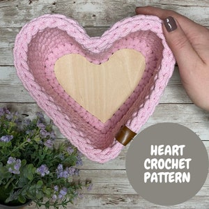 May include: A pink crocheted heart-shaped basket with a wooden heart insert. The basket has a brown leather strap. The text "HEART CROCHET PATTERN" is on a gray circle in the lower right corner of the image.