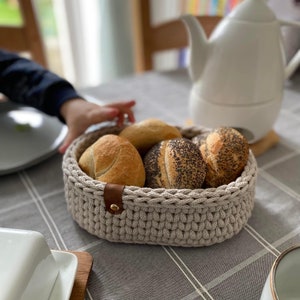 Może przedstawiać: Koszyk na chleb zrobiony na szydełku w kolorze beżowym z brązowym skórzanym uchwytem. Koszyk jest wypełniony sześcioma bułeczkami, niektóre z nich posypane są makiem.