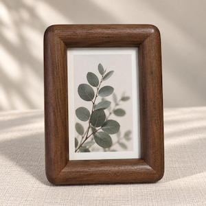 May include: A rectangular picture frame crafted from dark brown wood with a thick, rounded border. The frame holds a vertical print featuring a botanical illustration of a green leafy branch against a light, neutral-colored background.