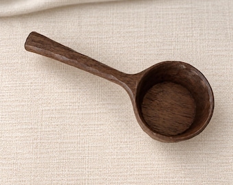 May include: A dark brown wooden scoop with a rounded bowl and a straight handle. The surface of the scoop features visible wood grain and a textured, hand-carved appearance. The scoop is positioned at an angle, showing the interior of the bowl.