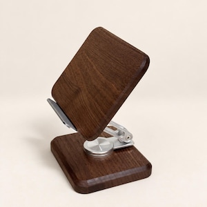 May include: A dark brown wooden phone stand featuring a rectangular base and a matching rectangular support panel. The two wooden components are connected by a silver-colored metal hinge mechanism that allows for adjustable positioning and tilting.
