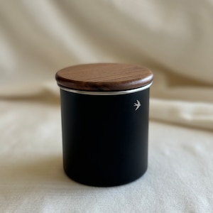 May include: Black canister with a wood lid and a small bird design. The canister is made of metal and has a silver rim.