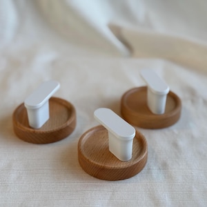 May include: Three wooden coasters with a white, rectangular handle attached to each. The coasters are made of light brown wood and have a circular shape. The handles are white and have a smooth, rectangular design.