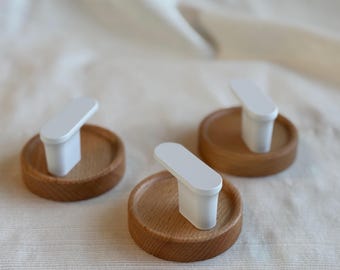 May include: Three wooden coasters with a white, rectangular handle attached to each. The coasters are made of light brown wood and have a circular shape. The handles are white and have a smooth, rectangular design.
