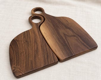 May include: Two dark brown wooden chopping boards with smooth surfaces and visible natural wood grain patterns. Each board features a rounded handle with a circular cutout at the top, designed for hanging or carrying the boards with ease.