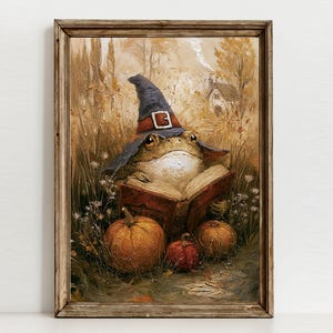 Op de afbeelding: Een ingelijste kunstdruk met een olieverfschilderij van een kikker die een donkerblauwe puntmuts met een gesp draagt en in een open boek leest. De scène speelt zich af in een grasveld met drie oranje pompoenen en een huisje op de achtergrond.