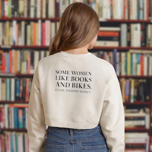 Puede incluir: Sudadera corta color crema con el texto "SOME WOMEN LIKE BOOKS AND BIKES. IT'S ME, I'M SOME WOMEN." La sudadera se combina con vaqueros azules. El fondo es una estantería borrosa.