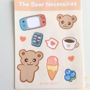 May include: A sheet of stickers featuring a brown bear, a video game console, a phone, a coffee cup, ice cream, blueberries, and hearts. The sheet is titled "The Bear Necessities" and has the artist's name "@anipangart" at the bottom.