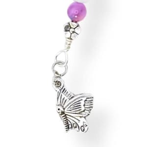 Puede incluir: Un dije de mariposa plateada cuelga de una pulsera de cuentas. La pulsera presenta cuentas blancas y moradas, con un adorno floral plateado. El dije de mariposa tiene alas grabadas.