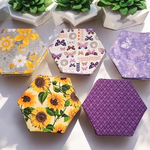 May include: A collection of six hexagonal fabric boxes with various floral and patterned designs. The patterns include sunflowers, butterflies, and abstract floral motifs in yellow, purple, and gray. The boxes are arranged on a white surface.