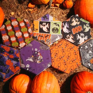 May include: A collection of Halloween-themed fabric face masks with various patterns, including pumpkins, ghosts, skeletons, and spiderwebs. The masks are arranged around several orange pumpkins on a bed of straw.