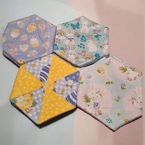 May include: A collection of hexagonal fabric coasters with Easter-themed patterns. Designs include bunnies, eggs, chicks, and floral motifs in pastel colors like blue, yellow, and green. These coasters are ideal for spring or Easter decor.