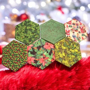 May include: A collection of six hexagonal fabric coasters with various Christmas-themed patterns. The coasters feature designs of holly, poinsettias, and festive figures. They rest on a vibrant red, fuzzy surface, with a blurred background of Christmas lights.