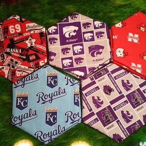 May include: Hexagon-shaped fabric items with sports team logos and text. Designs include "Huskers" in red, "K-State" in purple and gray, and "Royals" in blue. The items are arranged on a green surface.