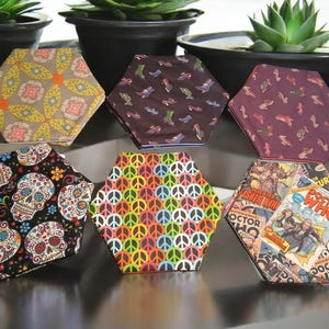 May include: A collection of six hexagonal fabric coasters with various patterns. Designs include floral, shoe prints, sugar skulls, peace signs, and comic book covers. The coasters are arranged on a reflective surface.