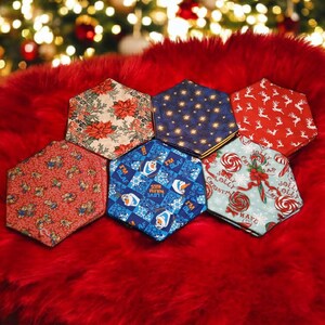 May include: A collection of hexagonal fabric coasters with various holiday-themed patterns, including poinsettias, reindeer, and snowflakes. The coasters are arranged on a red, fluffy surface, with a blurred Christmas tree in the background.