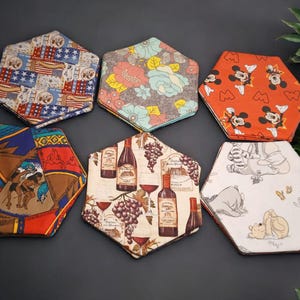 May include: Six hexagonal fabric coasters with various patterns. Designs include an American flag, floral, Mickey Mouse, equestrian, wine bottles and grapes, and Winnie the Pooh. The coasters are arranged on a dark surface.