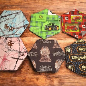 May include: Six hexagonal fabric coasters with various patterns. Designs include camouflage, John Deere tractor, red tractor on plaid, pink camouflage, "Game of Thrones" logo, and "Harry Potter" logo.