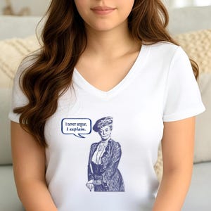 Op de afbeelding: Wit T-shirt met V-hals en een blauwe en witte illustratie van een vrouw met een hoed en een spraakballon die zegt "Ik discussieer nooit, ik leg uit."