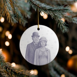 May include: A round, white Christmas ornament with a drawing of a man and woman in vintage attire. The couple is depicted in a grayscale illustration, with the man wearing a bowler hat and suit. The woman wears a hat and coat. Snowflakes are in the background.