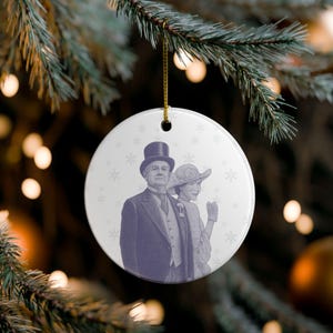 May include: A round, white Christmas ornament with a vintage illustration of a man and woman in formal attire. The couple is depicted in a purple hue, with a top hat and a decorated hat. Snowflake patterns are scattered across the ornament.