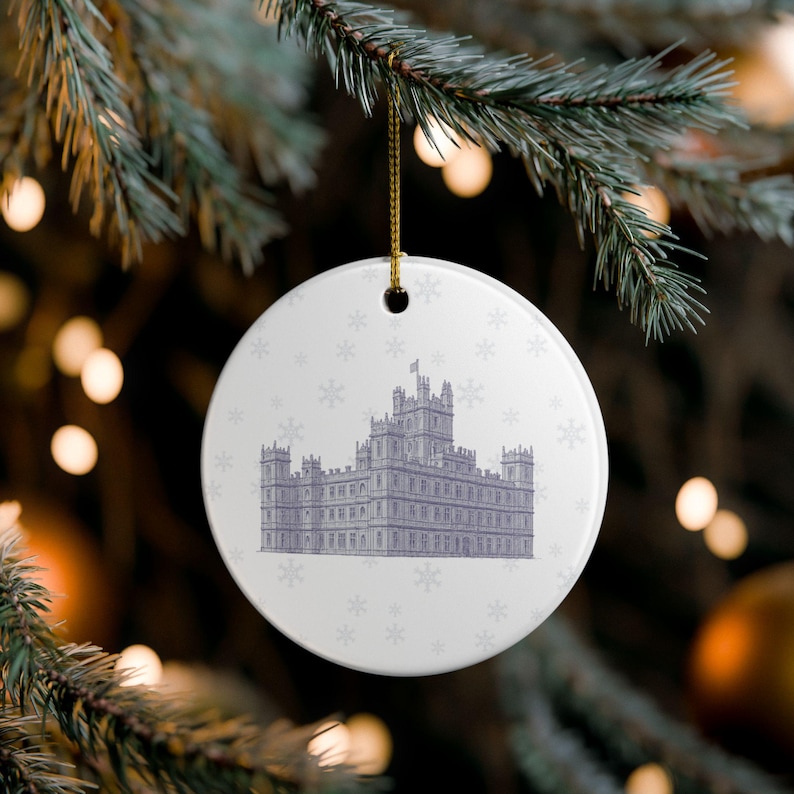 May include: White ceramic Christmas ornament with a detailed illustration of a building in shades of gray. The ornament has a gold string for hanging and is decorated with small snowflakes. The ornament is hanging from a green pine branch.