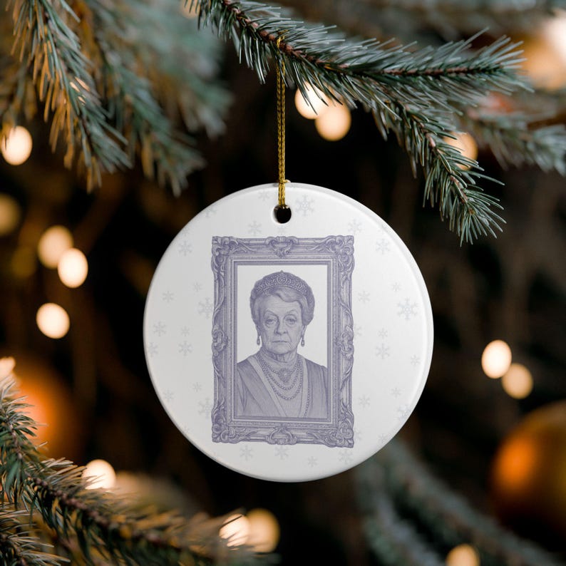 May include: A white, round Christmas ornament featuring a portrait of a woman in a decorative frame. The portrait is in shades of purple and gray, with snowflake accents around the edge. A gold string is attached for hanging.