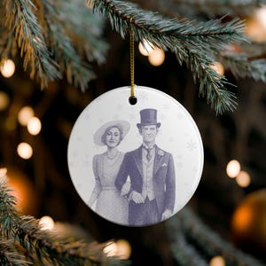 May include: A round, white Christmas ornament with a vintage illustration of a man and woman in formal attire. The man wears a top hat and suit, while the woman wears a hat and dress. The ornament has a gold string for hanging.