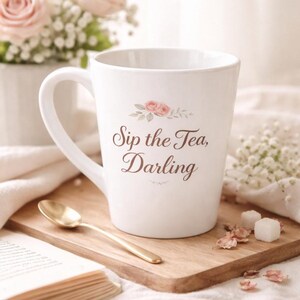 May include: White ceramic mug with the text "Sip the Tea, Darling" and a floral design. A gold spoon rests on a wooden board next to the mug. The mug is part of a tea set.