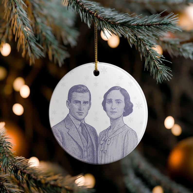 May include: A round, white Christmas ornament with a vintage illustration of a man and woman in a purple-grey hue. The ornament is decorated with small snowflake patterns and hangs from a gold string, perfect for holiday decor.
