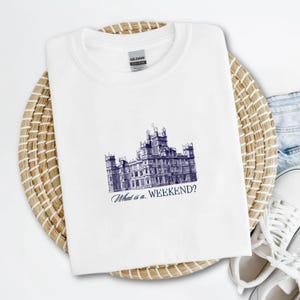 May include: White t-shirt with a blue and white illustration of a large house with the text "What is a... WEEKEND?"