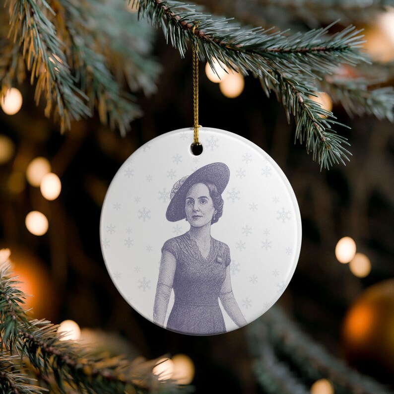 May include: A round, white Christmas ornament with a portrait of a woman in a hat and dress. The image is in shades of purple and gray, with snowflake accents. A gold string is attached for hanging.