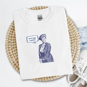 Può includere: Maglietta bianca con una stampa blu di una donna con un cappello e un fumetto che dice "Non discuto mai, spiego."