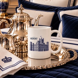 May include: A white ceramic mug featuring a blue illustration of a large castle-like building and the text "What is a WEEKEND?" printed in blue. The mug has a standard handle and sits on a metallic tray next to a silver teapot and creamer.