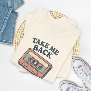 Puede incluir: Camiseta color crema con el texto "TAKE ME BACK" y un gráfico de cinta de cassette retro en tonos rojos, turquesa y marrón. La camiseta está doblada sobre una cesta tejida, con vaqueros azules y zapatillas blancas.
