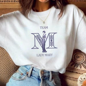 May include: White t-shirt with a graphic of a woman standing inside the letter "M" with the text "TEAM" above and "LADY MARY" below.