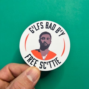 Puede incluir: Una pegatina redonda blanca con texto negro que dice "G'LFS BAD B'Y FREE SC"TTIE" y una imagen en blanco y negro de una persona con un mono naranja.