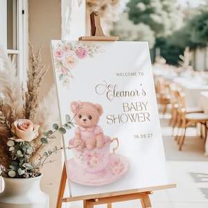 Puede incluir: Un letrero blanco sobre un caballete con las palabras "Welcome to Eleanor's Baby Shower" en letras doradas. Un osito de peluche con un vestido rosa está sentado en una taza de té rosa. Acentos florales en rosa y melocotón.