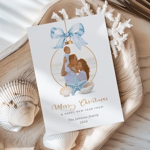 May include: A Christmas card with a family photo inside a gold bauble shape, decorated with a blue bow. The card reads "Merry Christmas & Happy New Year from The Johnson family 2026". The card is on a wooden tray with seashells and coral.