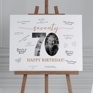 May include: A white sign on a wooden easel, celebrating a 70th birthday. The sign features the word "seventy" in gold script, with two black and white photos. The words "Happy Birthday!" are also in gold.