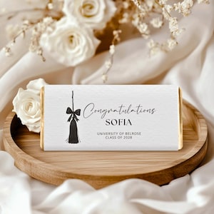 Op de afbeelding: Een witte chocoladereep met gouden randen, met de tekst "Congratulations SOFIA" en "UNIVERSITY OF BELROSE CLASS OF 2028". Een zwart kwastje en strik ontwerp staat boven de tekst. De reep ligt op een houten dienblad.
