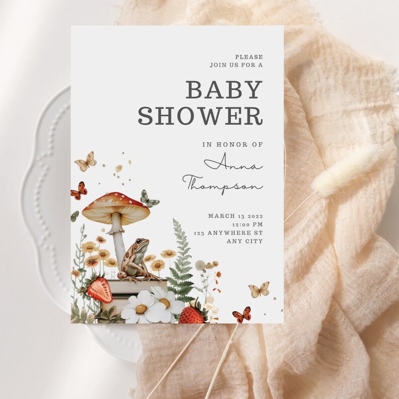 May include: A baby shower invitation with a whimsical woodland theme. The invitation features a mushroom with a frog sitting on top, surrounded by flowers, butterflies, and strawberries. The text on the invitation reads "Please join us for a baby shower in honor of Anna Thompson, March 13, 2022, 12:00 PM, 123 Anywhere St, Any City."