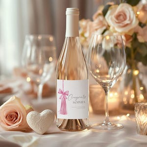 Peut inclure: Une bouteille de vin blanc avec un pompon et un nœud rose sur l'étiquette. L'étiquette indique "Congrats Sophia".