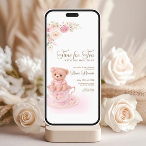 Puede incluir: Un teléfono inteligente muestra una invitación para un baby shower. La invitación presenta un osito de peluche en una taza de té, detalles florales y el texto "Time for Tea". La invitación incluye detalles para el baby shower de Anna Brown, incluyendo la fecha, hora y lugar.