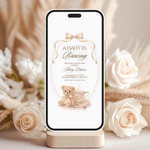 May include: A smartphone displays a baby shower invitation. The design features the text "A BABY IS Brewing" with a teddy bear and a cup of coffee illustration. The invitation includes the honoree's name, date, time, and location. The phone rests on a light-colored stand, surrounded by white roses.