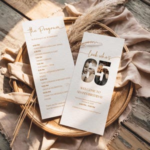 May include: Two cream-colored event programs with gold text, one titled "The Program" and the other "look who's 65". The programs are on a wooden tray with dried plants and a beige cloth. The program details a birthday celebration.