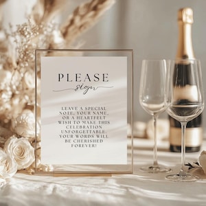 May include: A gold-framed sign with the words "PLEASE sign" in elegant script. The sign encourages guests to leave notes for a celebration. Two wine glasses and a champagne bottle are in the background, suggesting a wedding or party.