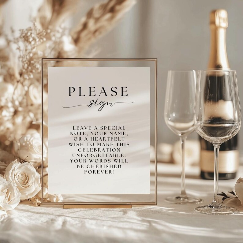 May include: A gold-framed sign with the words "PLEASE sign" in elegant script. The sign encourages guests to leave notes for a celebration. Two wine glasses and a champagne bottle are in the background, suggesting a wedding or party.