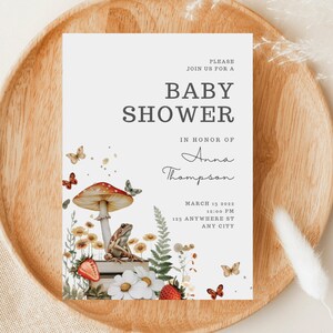 May include: A baby shower invitation with a whimsical woodland theme. The invitation features a red and white mushroom, a frog sitting on a stack of books, and butterflies. The text on the invitation reads "Please join us for a baby shower in honor of Anna Thompson, March 13, 2022, 12:00 PM, 123 Anywhere St, Any City."